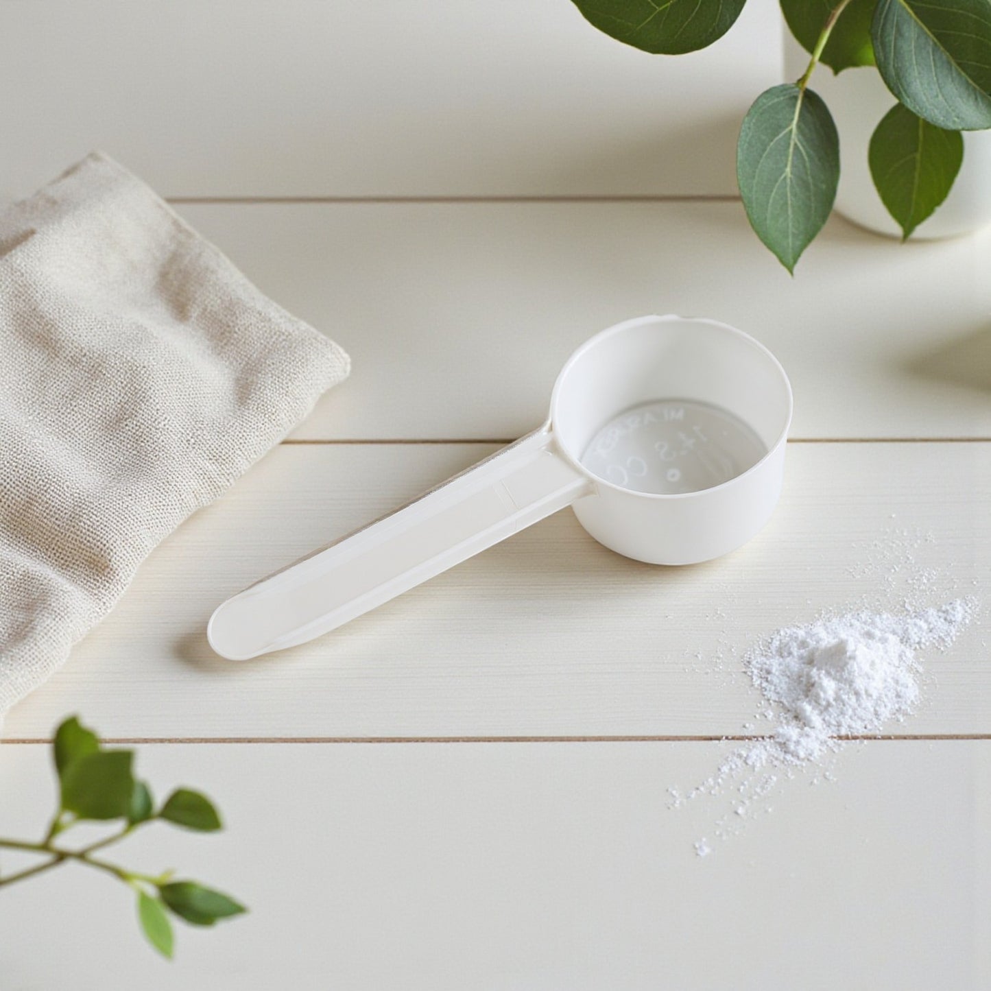 White measuring scoop with laundry powder on a light wooden surface with green leaves.