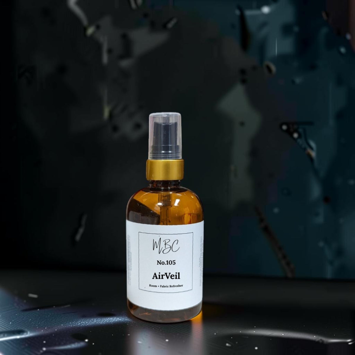 A small amber glass spray bottle labeled 'MBC No.105 AirVeil' with a white label and gold collar, standing on a dark reflective surface against a dark abstract background.