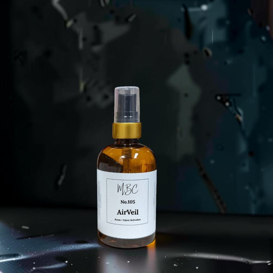 A small amber glass spray bottle labeled 'MBC No.105 AirVeil' with a white label and gold collar, standing on a dark reflective surface against a dark abstract background.