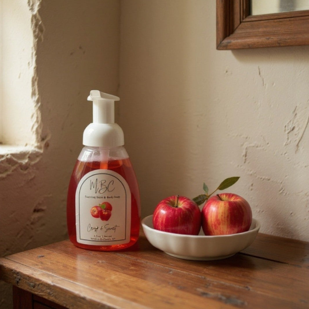 Bottle of soap with a label and apples on a wooden surface near a window.