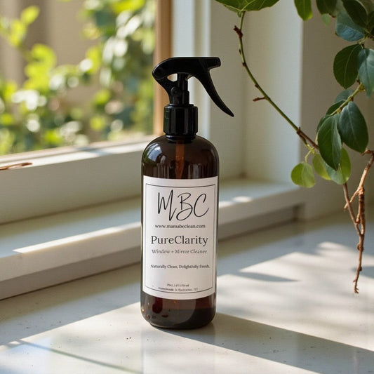 Mama Be Clean PureClarity glass and mirror cleaner in an amber spray bottle on a bright windowsill with greenery.